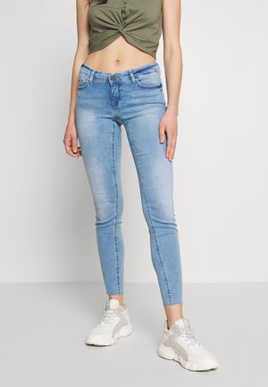 Femme portant un jean skinny bleu clair, des baskets blanches épaisses et un crop top torsadé vert olive, debout devant un fond blanc uni.