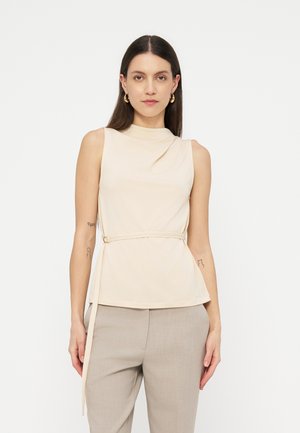 Mujer con top sin mangas beige con cinturón lateral y pantalones taupe claros, de pie contra un fondo blanco liso