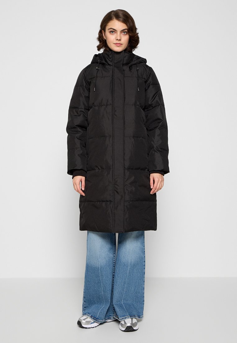 Puffermantel aus schwarzem Stoff mit Reißverschluss vorne, seitlichen Taschen und verstellbarem Kapuzen; kombiniert mit hellen, weiten Jeans und Sneakers.