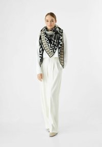 Jonge vrouw met een zwart, wit en beige geometrisch patroon sjaal over een witte trui en wide-leg witte broek, staande voor een effen achtergrond.