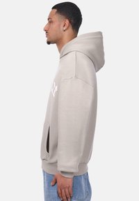 Hellbeige Kapuzenpullover mit geräumiger Kängurutasche, Zugbändern an der Kapuze und gerippten Bündchen. Auf der Vorderseite befindet sich weißer Drucktext.