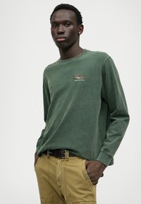 Grünes Langarm-T-Shirt mit Rundhalsausschnitt, ausgestattet mit einem kleinen gestickten Fischdesign und einem lockeren Schnitt; kombiniert mit khakifarbenen Hosen.