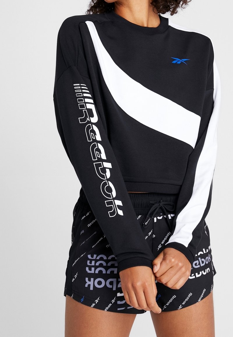 Sweat-shirt noir avec une bande diagonale blanche, présentant un logo Reebok blanc sur la manche. Associé à un short noir affichant un motif Reebok.