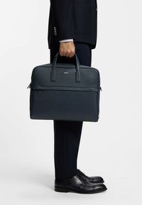 Ciemnoniebieska skórzana teczka z podwójnymi uchwytami i przednią kieszenią na suwak, trzymana przez osobę w garniturze i czarnych butach. Zawiera subtelne logo marki.