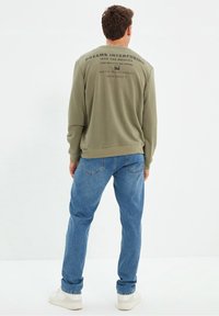 Felpa verde oliva con scritta sulla schiena, abbinata a jeans azzurri e sneakers bianche. Look casual, vestibilità rilassata con polsini a costine.