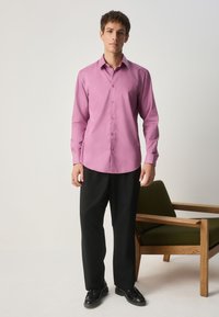 Hellrosa langärmliges Hemd mit Knöpfen und tailliertem Schnitt, kombiniert mit schwarzen Hosen und schwarzen Anzugschuhen, stehend vor einem grünen Stuhl.