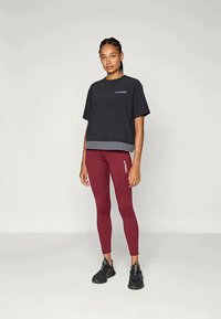 Čierne skrátené tričko s sivým lemovaním, v kombinácii s bordovými legínami s logom. Model má na sebe čierne športové topánky. Jednoduchý dizajn.