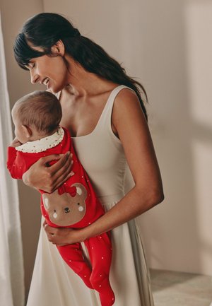 Salopetă roșie pentru bebeluși cu un design de urs și o fundiță, având buline albe. Adultă îmbrăcată într-o rochie de culoare deschisă, ținând bebelușul pe umăr.
