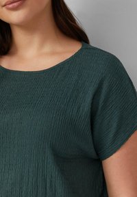 Teal gekleurde top met een subtiel kreukelontwerp, met een brede halslijn en korte mouwen. Gladde stof met een zachte glans.