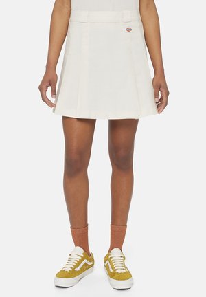 Femme portant une jupe blanche plissée avec un petit logo à la taille, associée à des chaussettes orange et des baskets jaune moutarde.