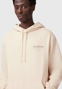 Felpa leggera color beige chiaro con cappuccio regolabile, tasca frontale e logo "ALLSAINTS" sul petto. Tessuto morbido con una finitura leggermente strutturata.