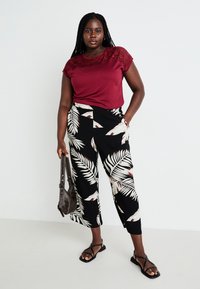 Burgundy t-shirt with lace detail at the shoulders, paired with black palm leaf-patterned pants. Model wears black flat sandals and holds a bag.