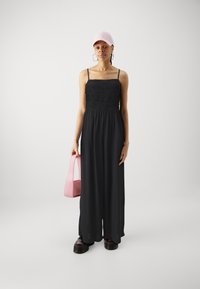 Svart maxi-kjole med smock-bodice, tynne stropper og løs passform. Kombinert med en lys rosa caps og en liten rosa håndveske. Tykk svart platåsko.