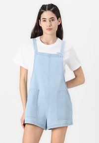 Világoskék farmer overall rövidnadrág széles pántokkal, két elülső zsebbel és laza szabással. Egyszerű fehér pólóval alatta.