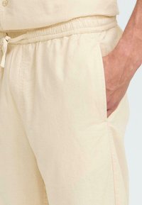 Lys beige bukser med elastisk talje, snor detalj og sidelommer, laget av et mykt, teksturert stoff.