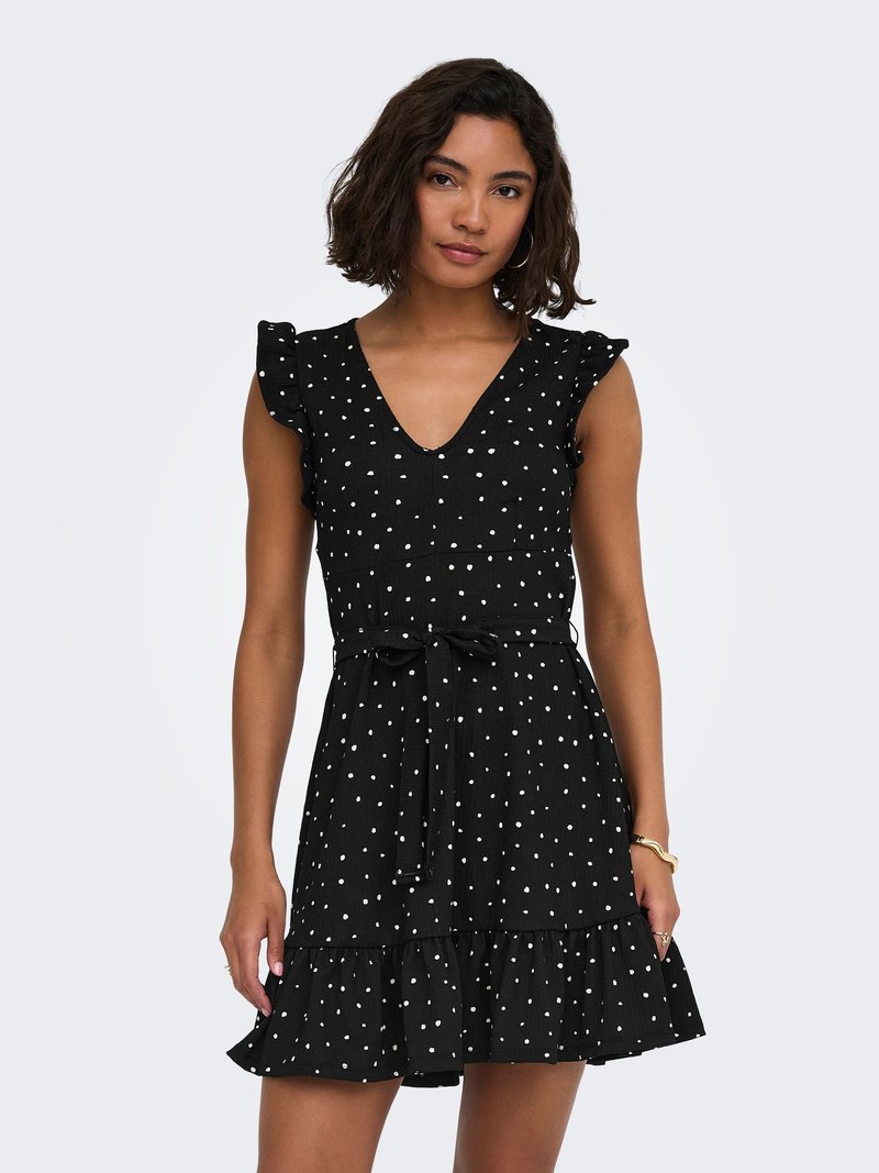 Femme portant une robe noire sans manches à pois blancs, manches à volants, ceinture nouée à la taille et ourlet à volants, debout devant un fond uni.