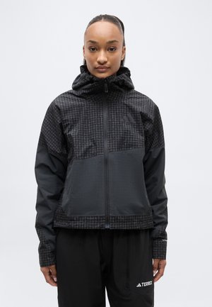 Nő fekete mintás kapucnis dzsekiben és fekete nadrágban, amelyen Terrex logó látható, egy egyszerű fehér háttér előtt áll.