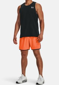 Svart ärmlös träningstopp; klarröda shorts med svart kant; person som bär vita sportskor, stående framför en neutral bakgrund.