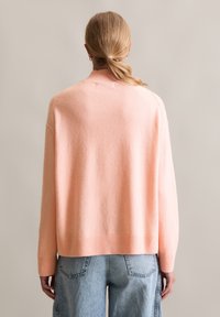 Maglione oversize rosa chiaro con polsini e orlo a coste, texture morbida e colletto a monte, indossato sopra jeans a vita alta blu chiaro.