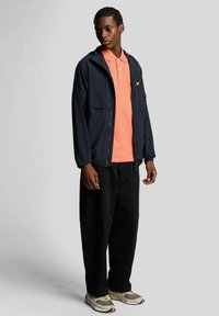 Marineblauw jack met rits en twee steekzakken aan de voorkant, gedragen over een lichtoranje poloshirt. Zwart broek met wijde pijpen en grijze sneakers maken de outfit compleet.