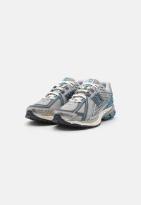 Pár šedých a tyrkysových běžeckých bot New Balance s bílými šňůrkami, síťovaným designem a polstrovanou podešví na bílém pozadí.