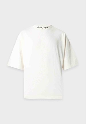 Homme debout portant un t-shirt blanc oversize et un pantalon marron ample avec des baskets blanches sur un fond clair uni.
