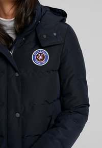 Doudoune bleu marine en tissu lisse, dotée d'une fermeture éclair et de boutons-pression, d'une capuche, et d'un logo rond brodé sur la poitrine.