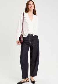 Blouse blanche avec un col à volants, manches longues ; associée à un jean en denim bleu foncé taille haute. Chaussures plates noires ; petit sac bandoulière marron.
