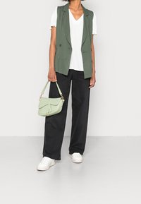 Zielona bezrękawnikowa marynarka na białej bluzce z dekoltem w serek, zestawiona z czarnymi spodniami o szerokich nogawkach i białymi trampkami, trzymająca jasnoszaroniebieską torebkę.