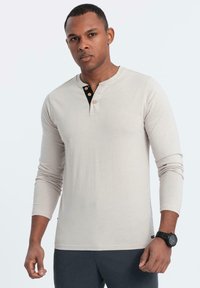 Lichtbeige langarm henley shirt met zwarte pat en drie knopen; soepele stof; strakke pasvorm; gecombineerd met donkere broek.