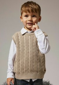 Chaleco de punto beige con diseño de cable sobre una camisa blanca con cuello, con costuras texturizadas, diseño sin mangas y un dobladillo que se sitúa por encima de la cintura.