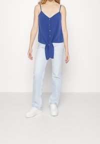 Niebieska, bezrękawnikowa bluzka z węzłem w talii i zapięciem na guziki z przodu. Połączona z jasnoniebieskimi, prostymi dżinsami i białymi trampkami.