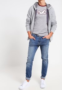 Grå zip-up hoodie över en grå t-shirt med en logotyp, blå slim-fit jeans och vita sneakers. Utstyrseln har en avslappnad design.