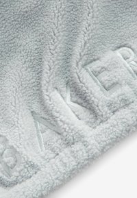 Ljusgrå fleecefilt med en texturerad, fluffig yta. Har upphöjda broderade bokstäver som ger detaljer och djup till designen.