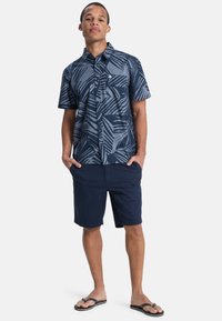 Marineblauw overhemd met korte mouwen en een bladerenpatroon, met knoopsluiting en zak; gecombineerd met marineblauwe shorts en slippers.