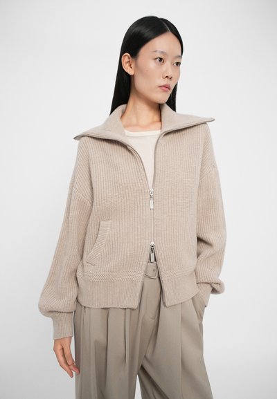 Beige glidelås-cardigan med høy krage, ribbestruktur, to sidelommer og voluminøse ermer, sammen med lyse bukser.