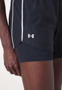 Närbild av en person som bär svarta Under Armour sportshorts i lager med vit kant på det yttersta lagret.