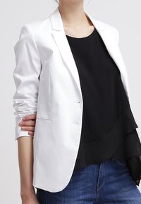 Blazer bianco con un solo bottone, spalle strutturate e due tasche frontali, indossato sopra una maglia nera con volant e jeans blu.