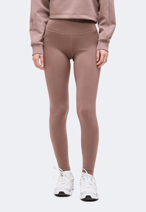 Persoon draagt mauve strakke legging, bijpassende cropped trui en witte sneakers, staat met handen ontspannen langs het lichaam.