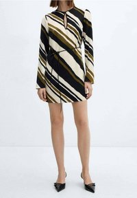 Frau trägt ein langärmliges, knielanges Kleid mit diagonalen schwarzen, weißen und braunen Streifen, tailliert mit einem dünnen schwarzen Gürtel.