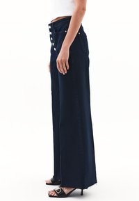 Szerokie granatowe dżinsy wykonane z denimu, z wysokim stanem, licznymi złotymi guzikami oraz postrzępionym wykończeniem na dole.