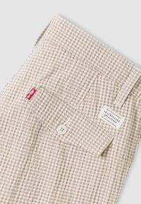 Ύφασμα με μπεζ και λευκό καρό σχέδιο, διπλωμένο με τσέπη που κλείνει με κουμπί. Διαθέτει ετικέτα Levi's και έχει λείο, υφασμάτινο φινίρισμα.