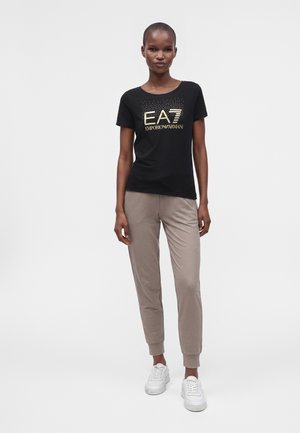 Femme portant un t-shirt noir avec le logo doré EA7 Emporio Armani, un pantalon de jogging taupe et des baskets blanches, debout devant un fond uni.