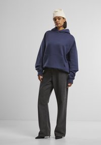 Overdimensjonert marineblå hettegenser med frontlomme, sammen med høyspent, løst sittende mørk grå jeans og en kremfarget strikket lue.