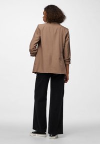 Mujer con cabello rizado que lleva una chaqueta marrón, pantalones anchos negros y zapatillas negras con blancas, de pie de espaldas sobre un fondo blanco.