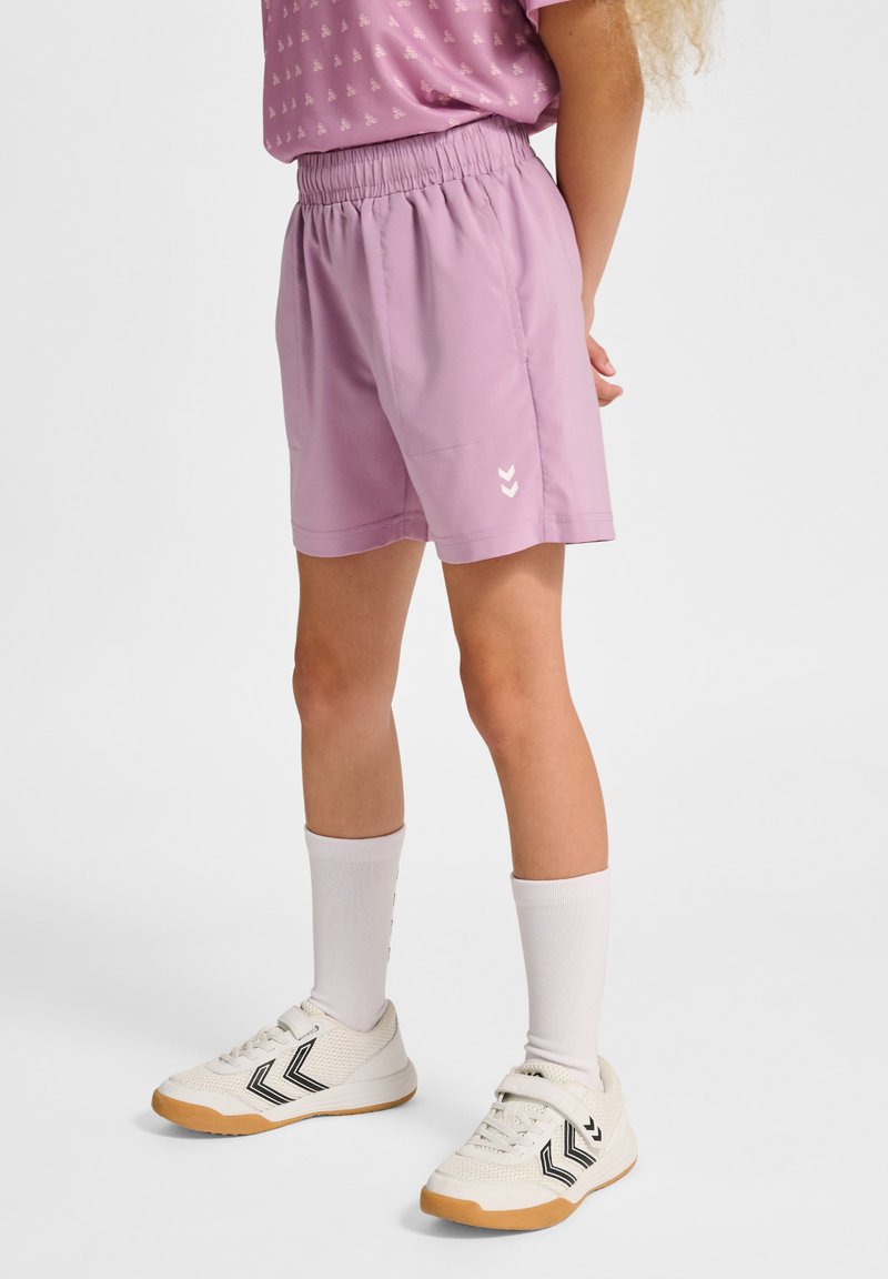 Lichtpaarse shorts van zachte stof met een elastische tailleband, voorzien van witte chevron-logo's aan de onderzijde links. Gestyled met witte sokken en sneakers.
