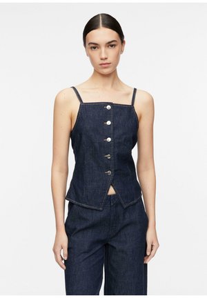 Młoda kobieta o ciemnych włosach, ubrana w ciemny jeansowy top bez rękawów zapinany na guziki oraz pasujące ciemne jeansowe spodnie na jednolitym tle.