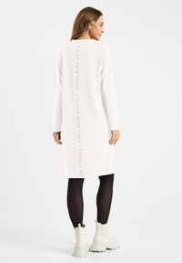 Witte sweatshirt jurk met lange mouwen, voorzien van een verticale roze logo afdruk op de achterkant. Gecombineerd met zwarte legging en witte laarzen.
