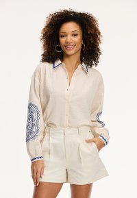 Camicia beige con bottoni, con accenti ricamati blu sul colletto e sulle maniche, abbinata a pantaloni corti beige a vita alta. Tessuto strutturato.