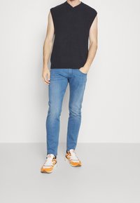 Mörkgrå ärmlös stickad topp, blå slim-fit jeans och sportiga vita sneakers med orange accenter, stående mot en neutral bakgrund.